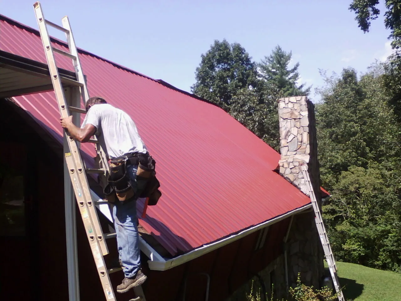 Roofer on a ladder beside a red metal roof and stone chimney — Soffit & Fascia Repair by Roofing Brothers in Payne